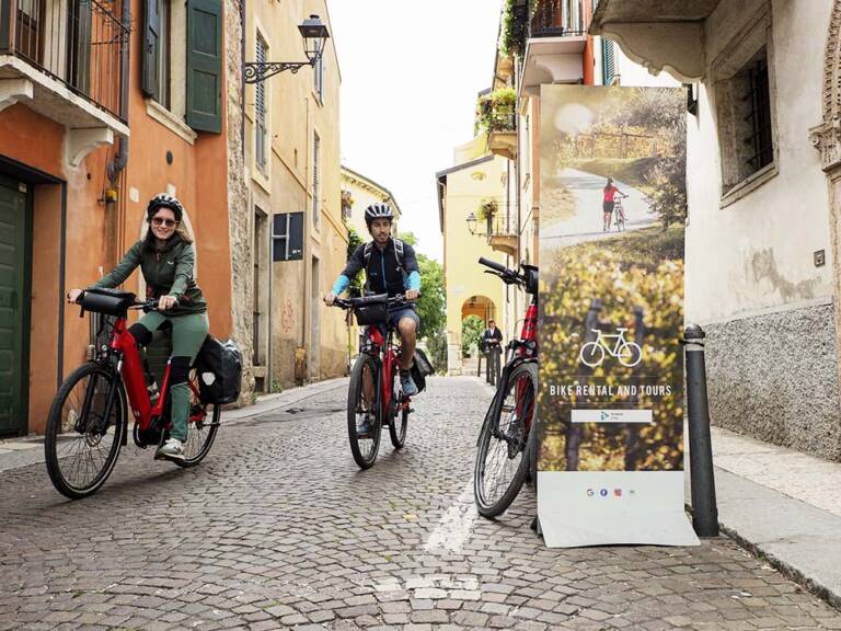Due ciclisti su e-bike da cicloturismo percorrono un vicolo acciottolato del centro storico di Verona, vicino all'entrata del noleggio bici Itinera Bike